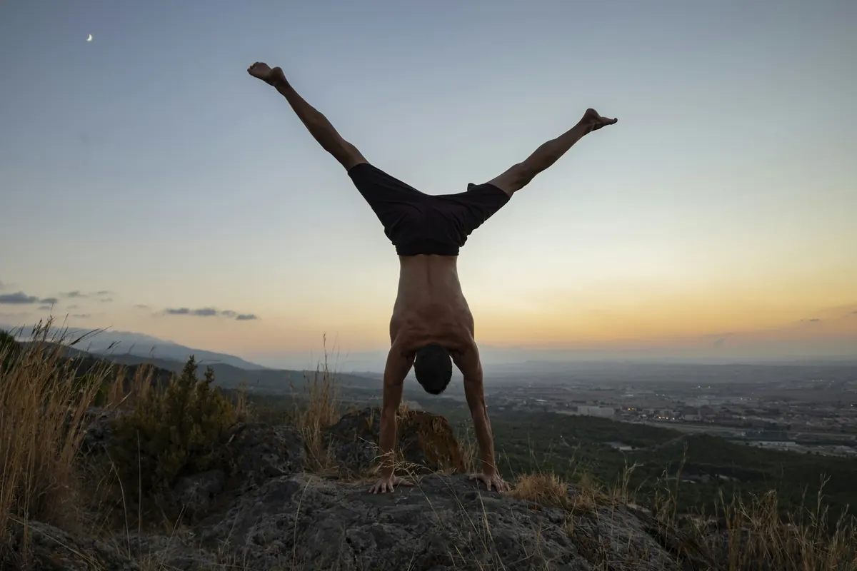An athlete holding a straddle handstand on a hilltop at sunset
